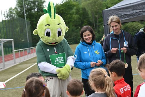 Person im grünen Jolinchen-Kostüm mit AOK-Logo neben Kristine Weinhold von der Stiftung Anstoß zum Leben und Nadine Dietrich vom Kreissportbund, die Kindern Medaillen überreichen auf Sportplatz